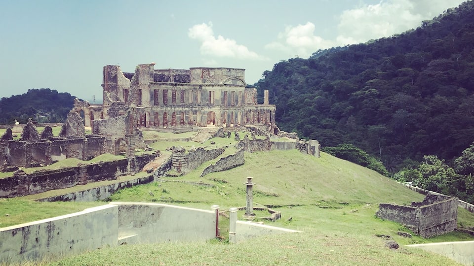 An ancient ruin with a large building and surrounding walls, set against a backdrop of a hill and clear sky.
