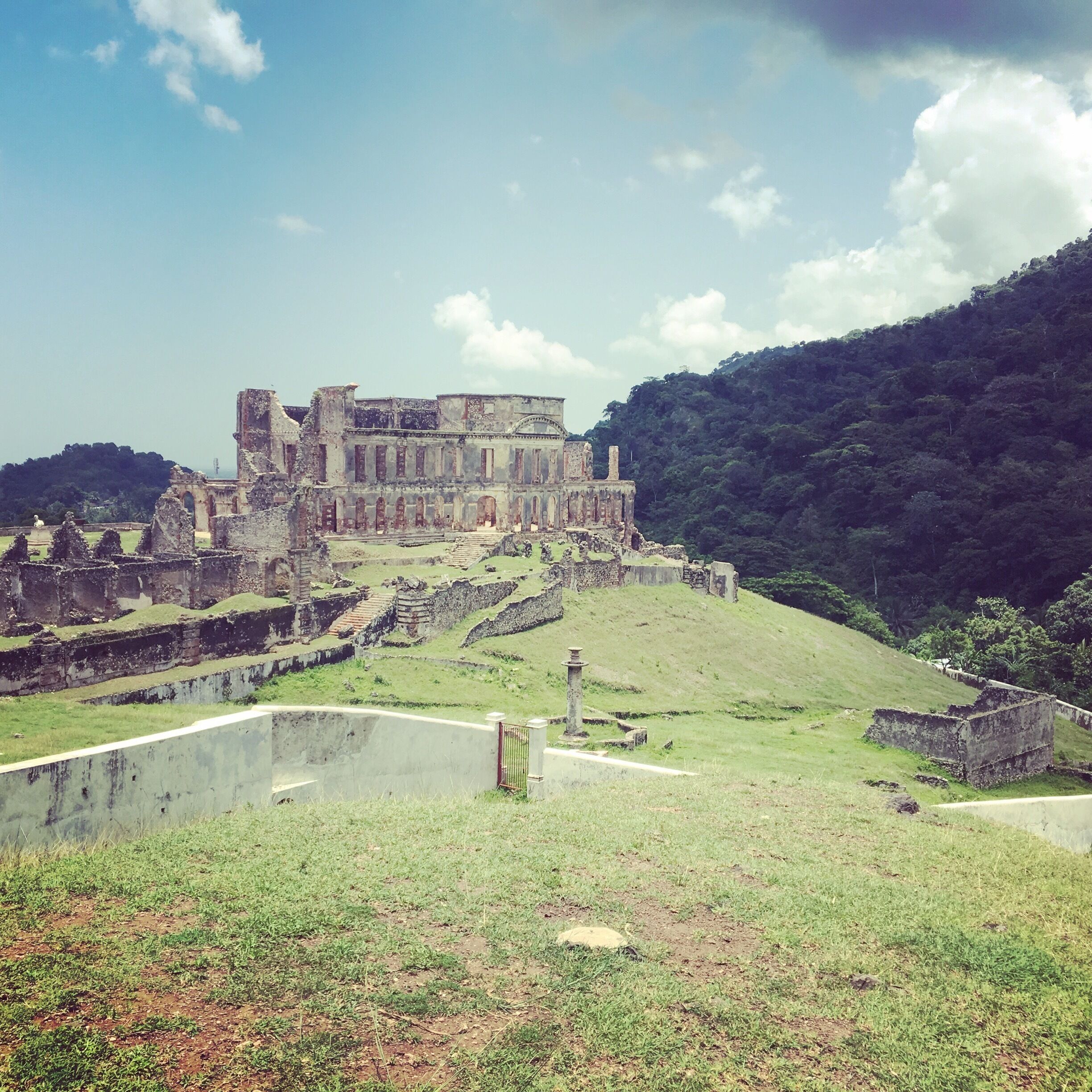 An ancient ruin with a large building and surrounding walls, set against a backdrop of a hill and clear sky.