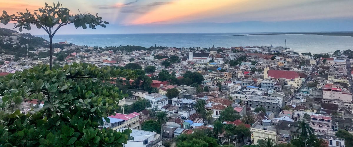 A coastal cityscape with a tree in the foreground, buildings, and a sunset over the sea.
