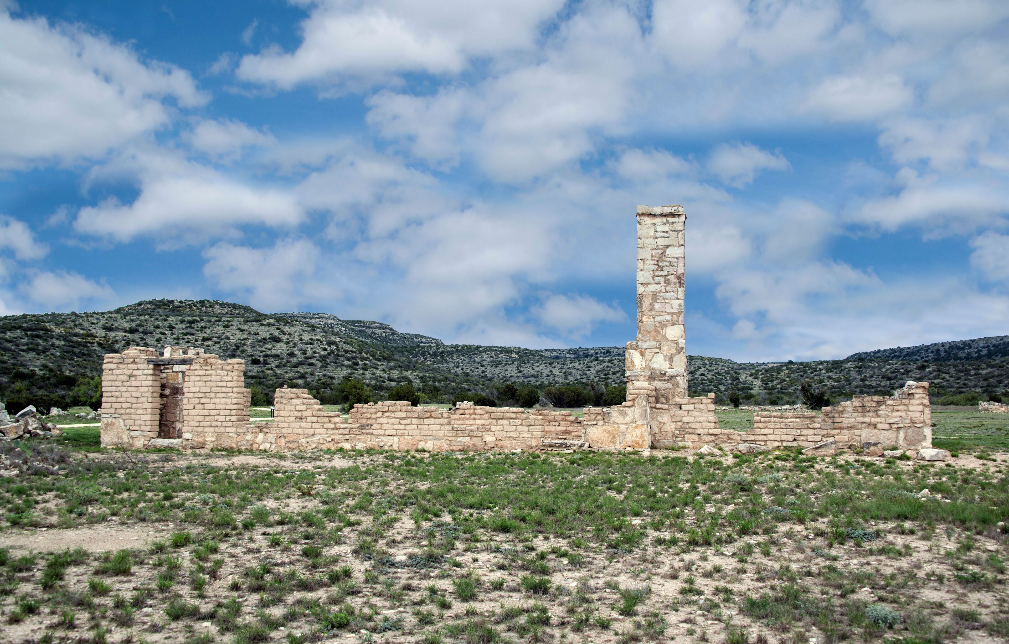 Ancient stone ruins with a tall column, surrounded by a grassy field and mountains in the background.
