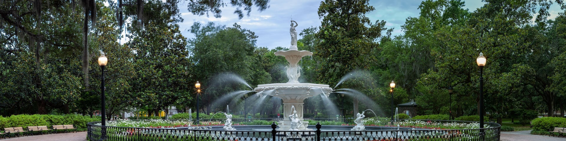 A circular fountain with statues and water jets, surrounded by a fence and benches, set in a park with trees and lamp posts.