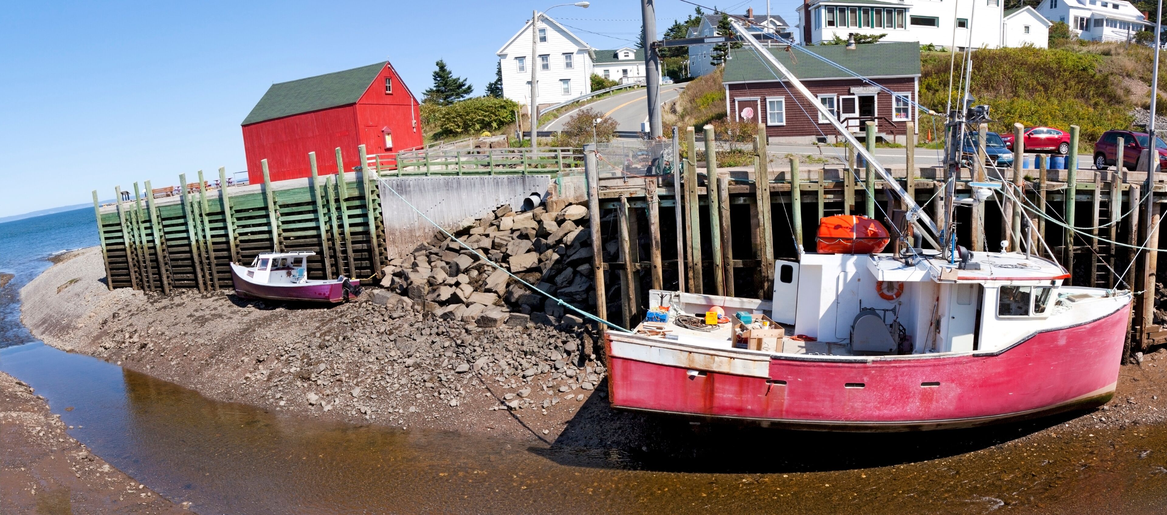 A red boat docked at a rocky shore with a red building and white houses in the background.