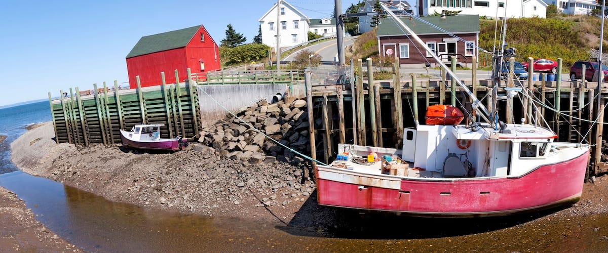 A red boat docked at a rocky shore with a red building and white houses in the background.