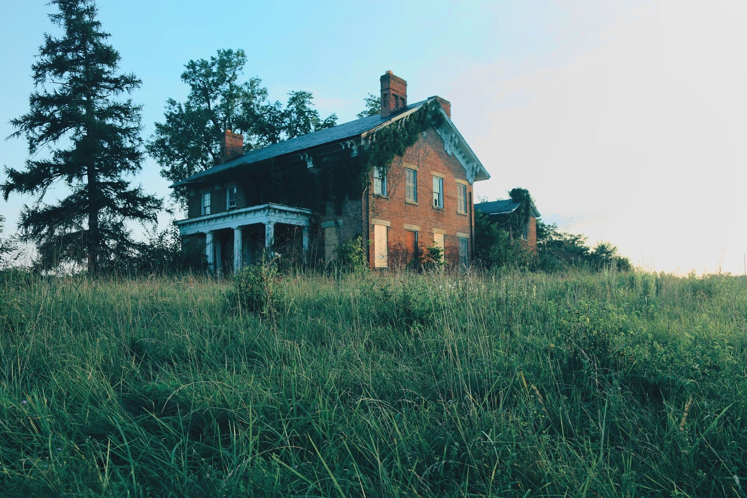 A large, abandoned brick house with a wrap-around porch, surrounded by tall grass and trees.
