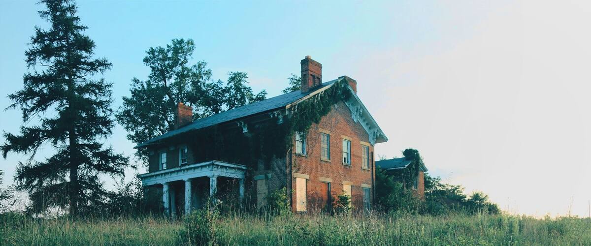 A large, abandoned brick house with a wrap-around porch, surrounded by tall grass and trees.