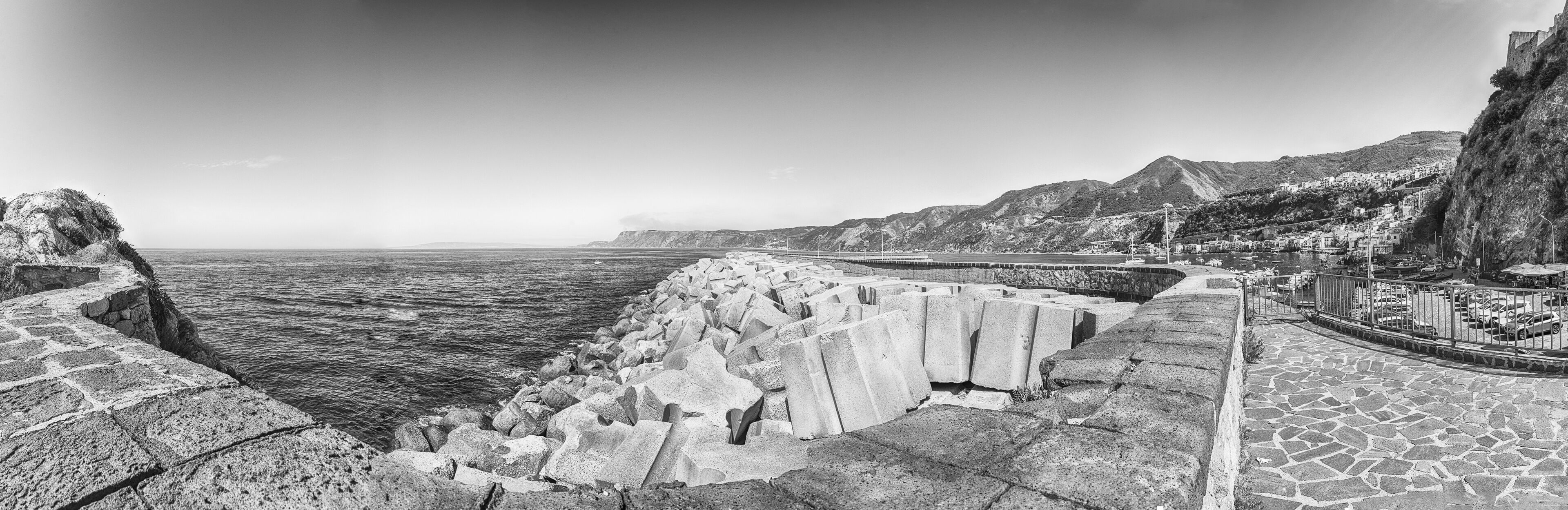 A coastal landscape with a stone sea wall, a paved promenade, and a mountainous backdrop.