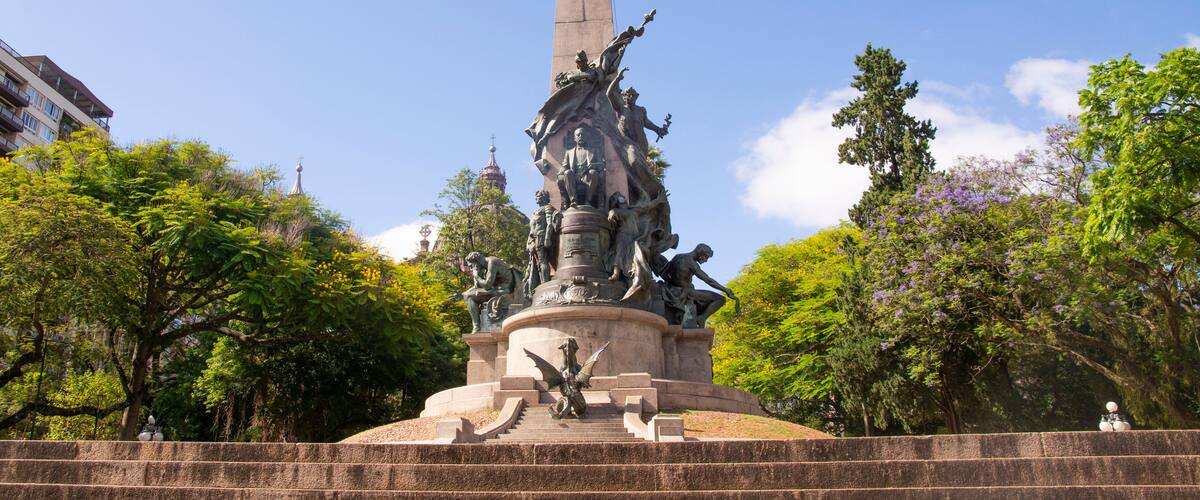 A tall monument with statues, surrounded by trees and a clear sky.