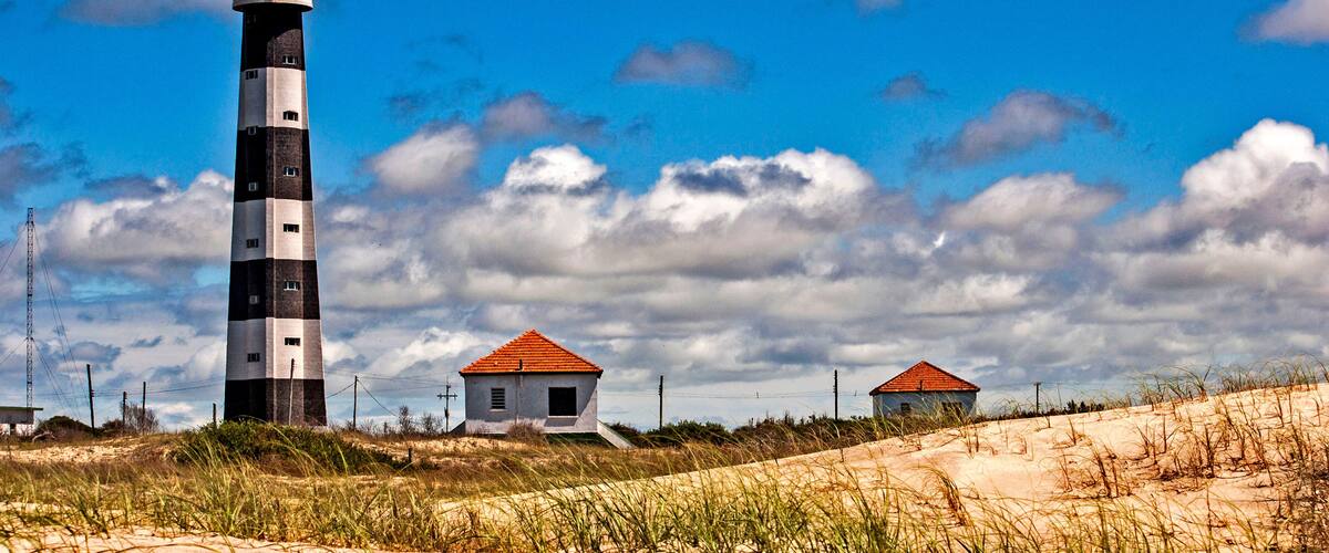 A lighthouse, two small buildings, and sand dunes under a partly cloudy sky.