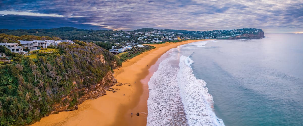 An aerial view of a coastal town with a long sandy beach, dense greenery, and a hilly backdrop.