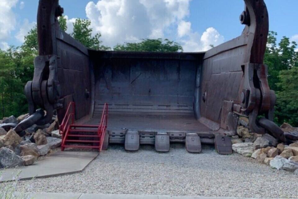 A large metal structure resembling a ship's bow, surrounded by gravel and plants, with a clear sky in the background.