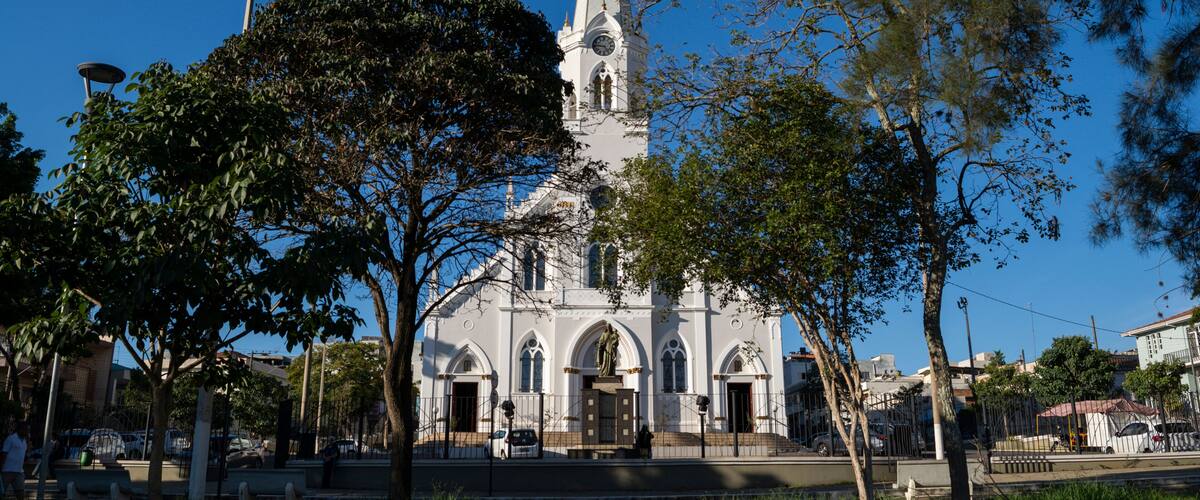 A white church with a tall steeple and a clock, surrounded by trees and a street.