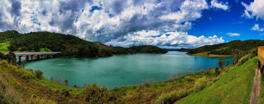 Panoramic photo of the Paraibuna River, in Sao Paulo, Brazil, running between hills on a sunny day with a blue sky and clouds