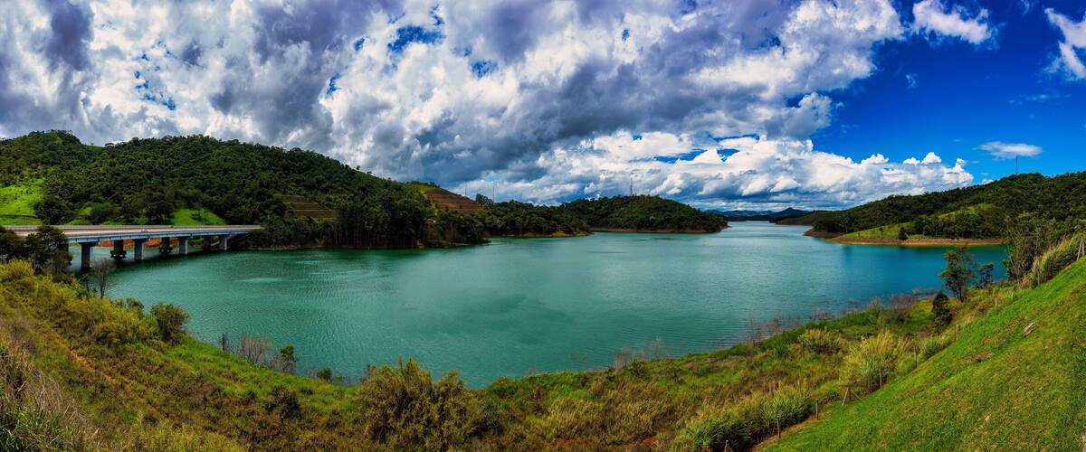 A river with a bridge, surrounded by lush green hills and a partly cloudy sky.