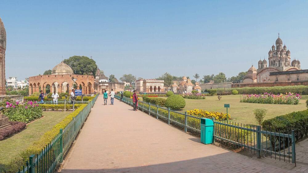 A historical site with a prominent dome structure, a pathway lined with greenery, and a large building with multiple domes in the background.
