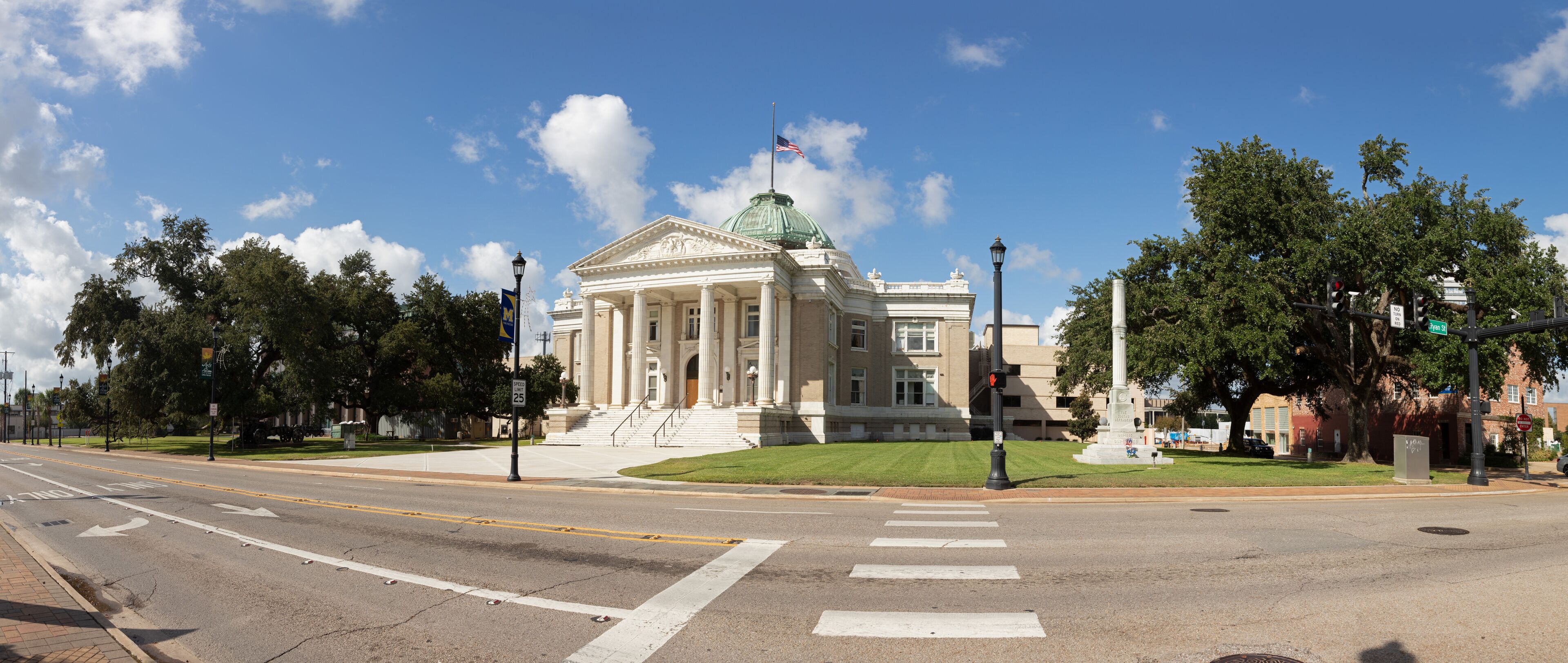 A large neoclassical building with a green dome and columns, located at a street intersection with traffic lights and a crosswalk.