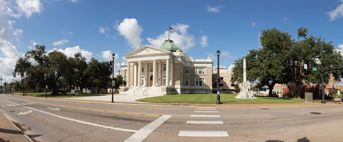 A large neoclassical building with a green dome and columns, located at a street intersection with traffic lights and a crosswalk.
