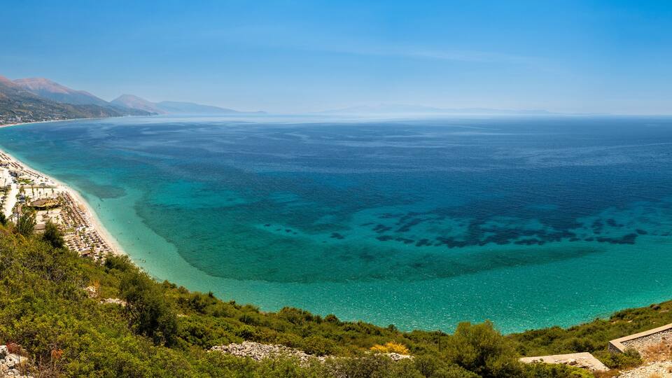 A coastal landscape with a clear blue sea, a sandy beach, and a mountainous backdrop.