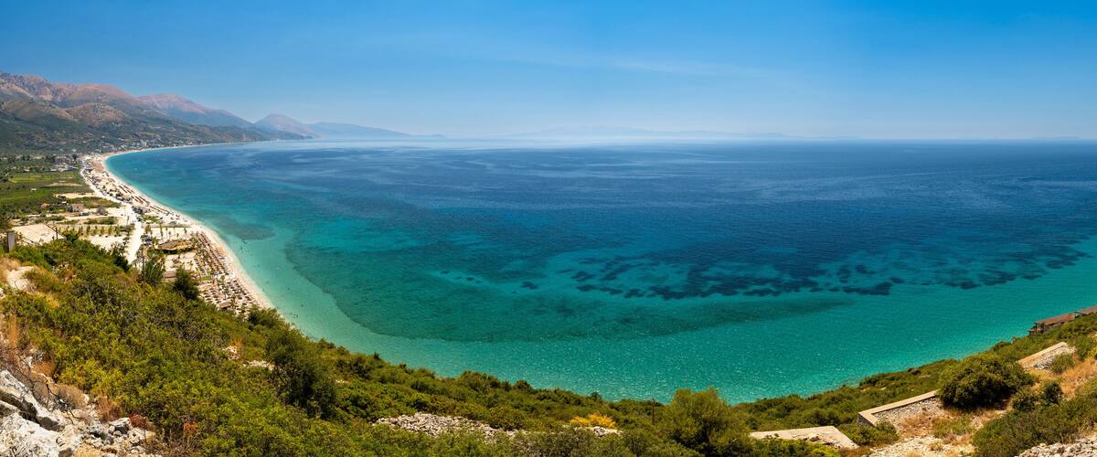 A coastal landscape with a clear blue sea, a sandy beach, and a mountainous backdrop.