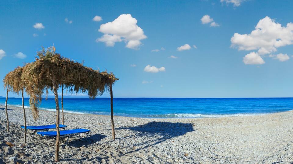 A pebble beach with a thatched umbrella and blue chairs, a clear blue sky, and calm sea.