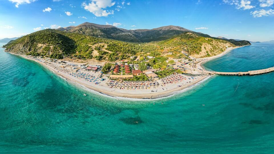 An aerial view of a coastal island with a beach, a pier, and mountainous terrain in the background.