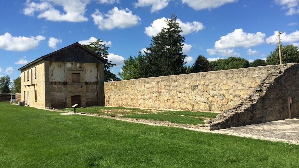A historic stone wall and a weathered building with a grassy field in front.