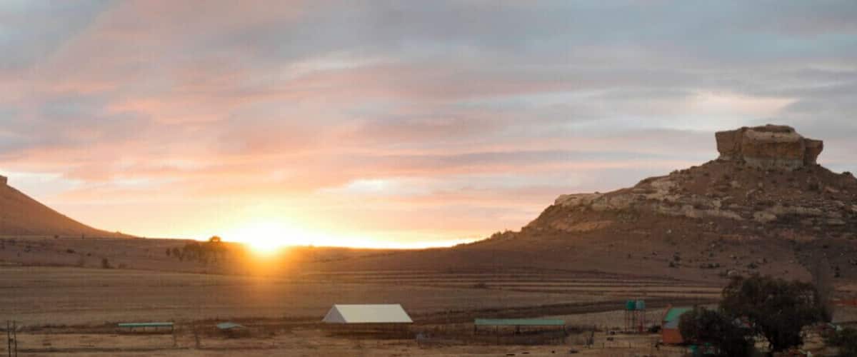 A sunset over a flat landscape with two prominent hills and a small building in the foreground.