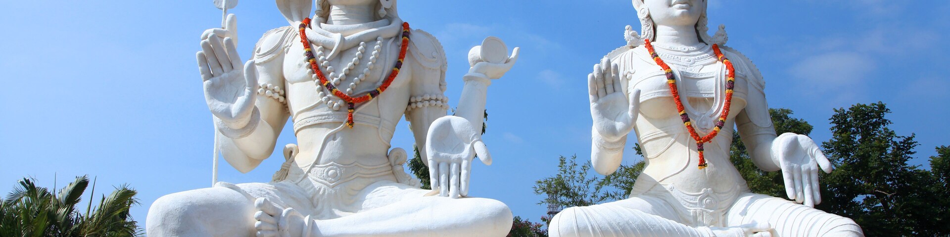 Duas grandes estátuas brancas de divindades, com tridentes, sentadas sobre uma rocha, com um céu azul límpido ao fundo.