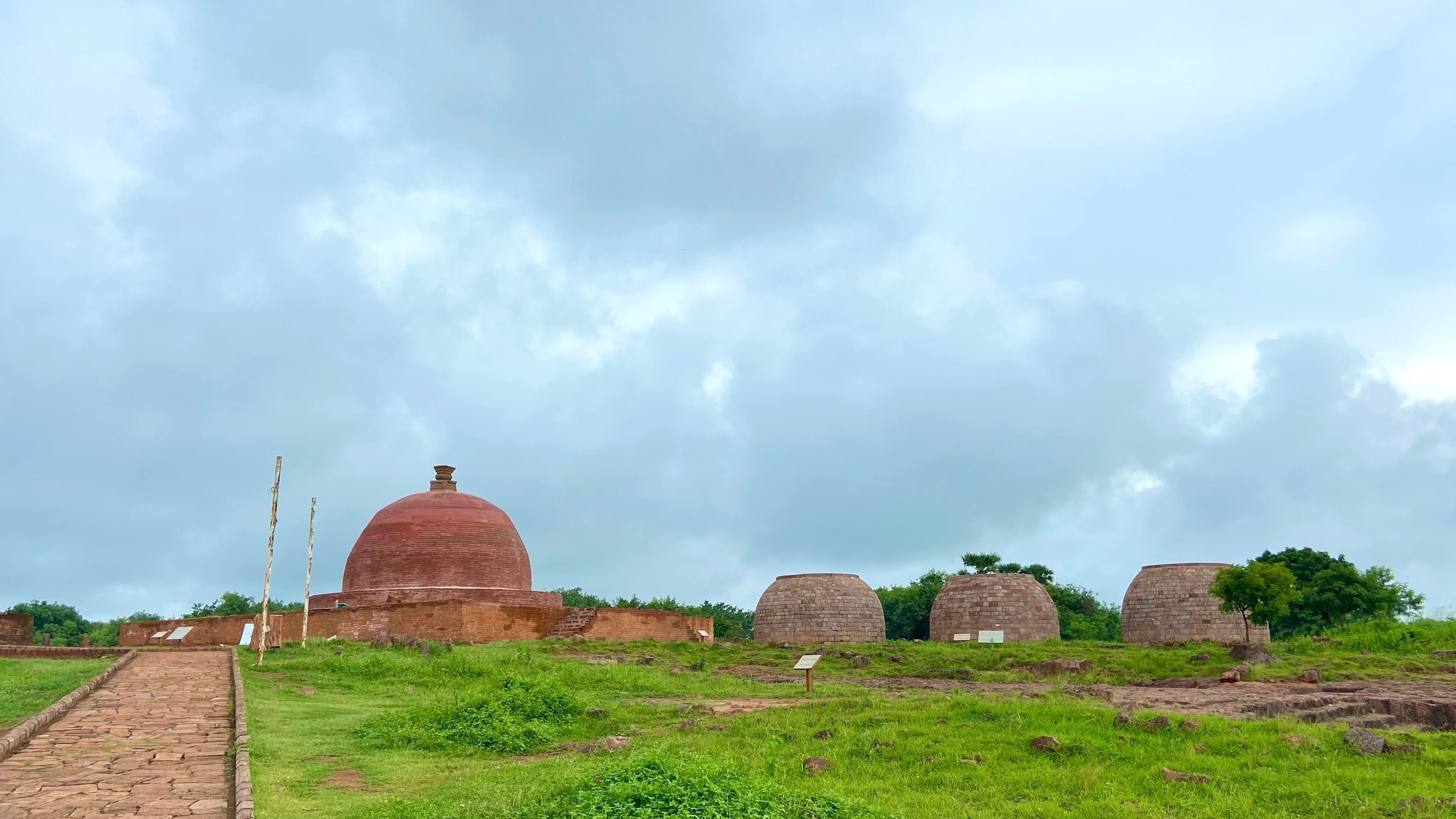 A pathway leading to ancient brick structures with domed roofs, surrounded by grassy fields and a cloudy sky.