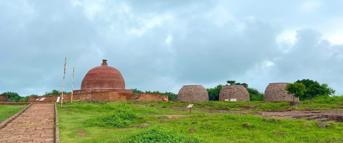 A pathway leading to ancient brick structures with domed roofs, surrounded by grassy fields and a cloudy sky.