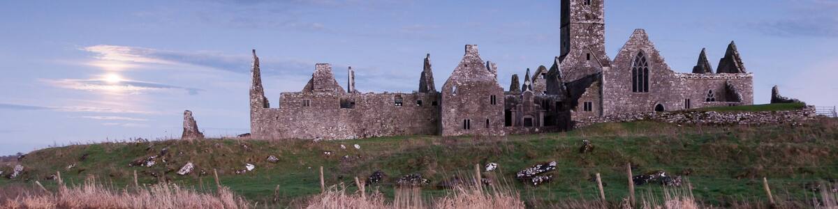 A historic stone castle with a prominent tower, situated on a grassy hill overlooking a calm body of water.