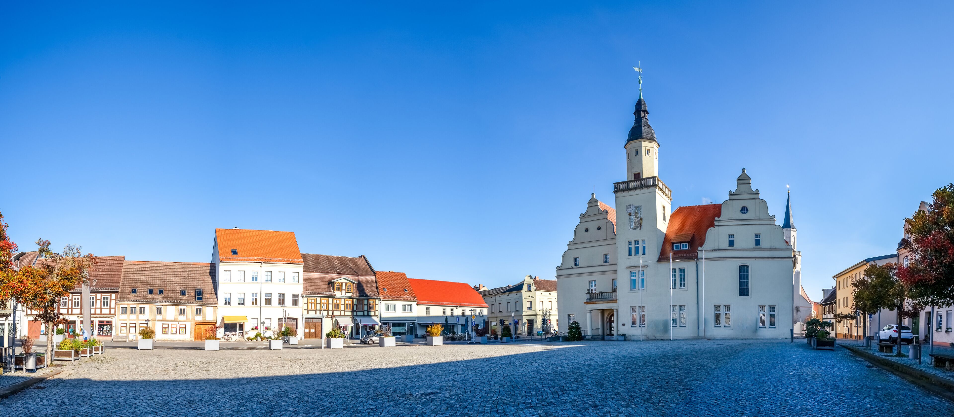 A cobblestone square with historic buildings, a prominent church with a tall spire, and a clear blue sky.