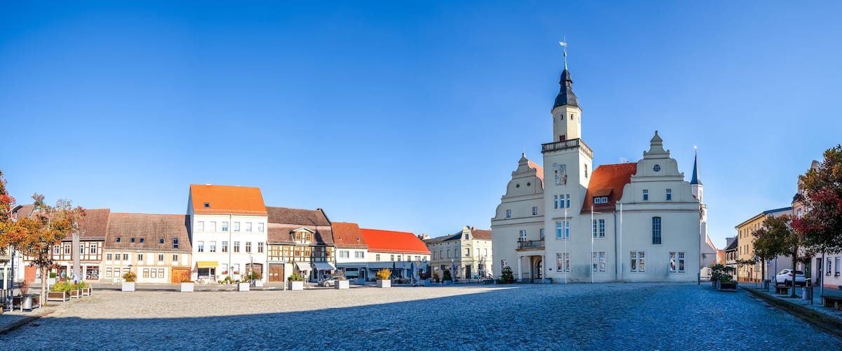 A cobblestone square with historic buildings, a prominent church with a tall spire, and a clear blue sky.