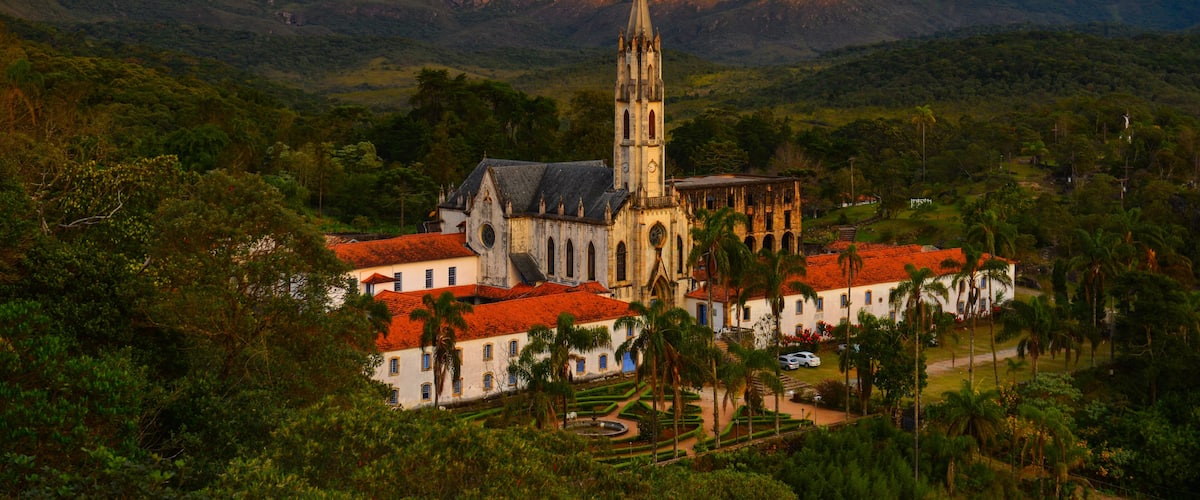 A church with a tall tower surrounded by lush greenery and palm trees, set against a backdrop of mountains.