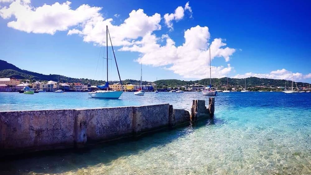 A coastal marina with boats, clear blue water, and a distant hilly landscape.