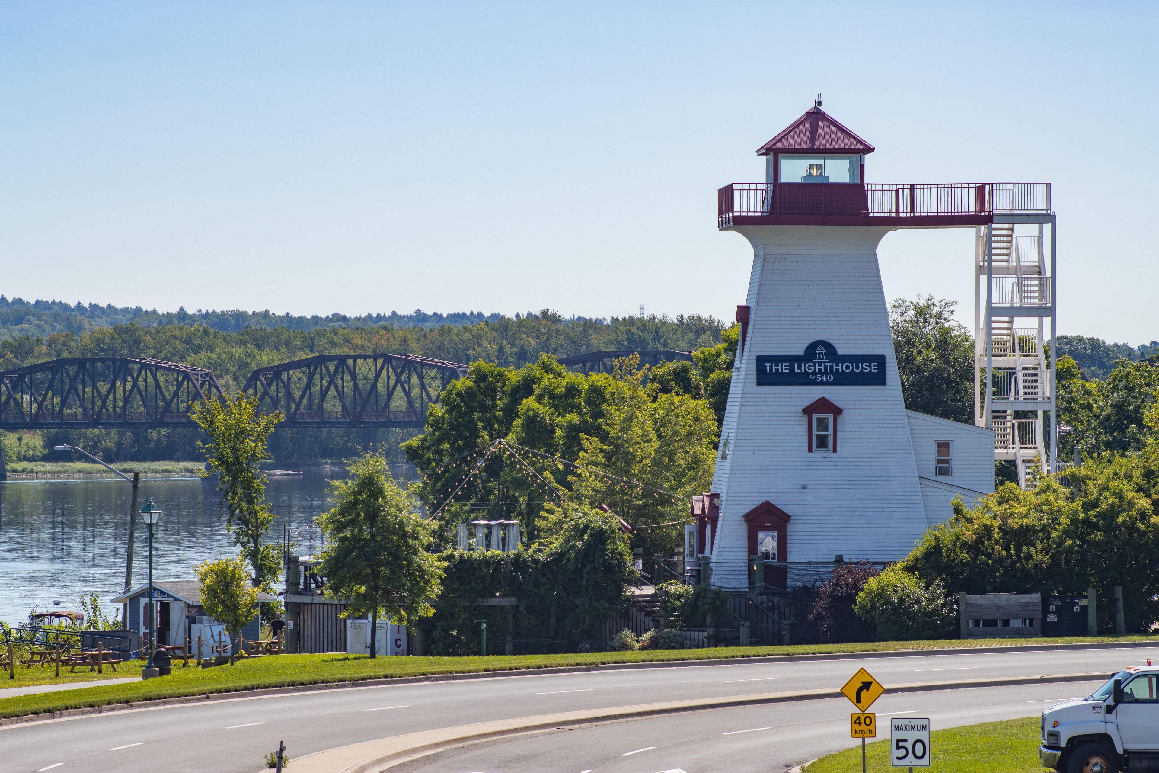 A lighthouse named "The Lighthouse" with a red roof and white walls, a bridge in the background, and a road with a speed limit sign.