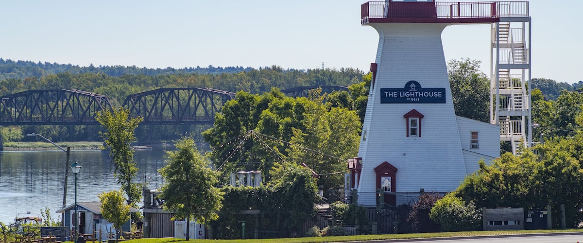 A lighthouse named "The Lighthouse" with a red roof and white walls, a bridge in the background, and a road with a speed limit sign.