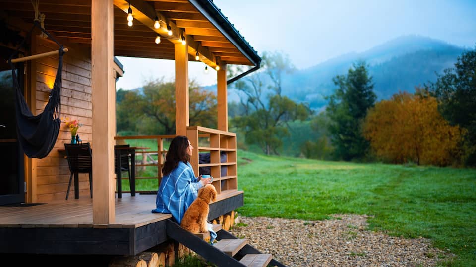 A wooden deck with a hammock, a woman, and a dog overlooking a grassy field and mountains.