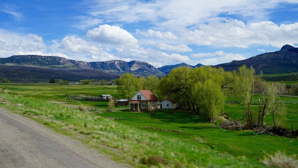 A rural landscape with a house, green fields, and mountains in the background.