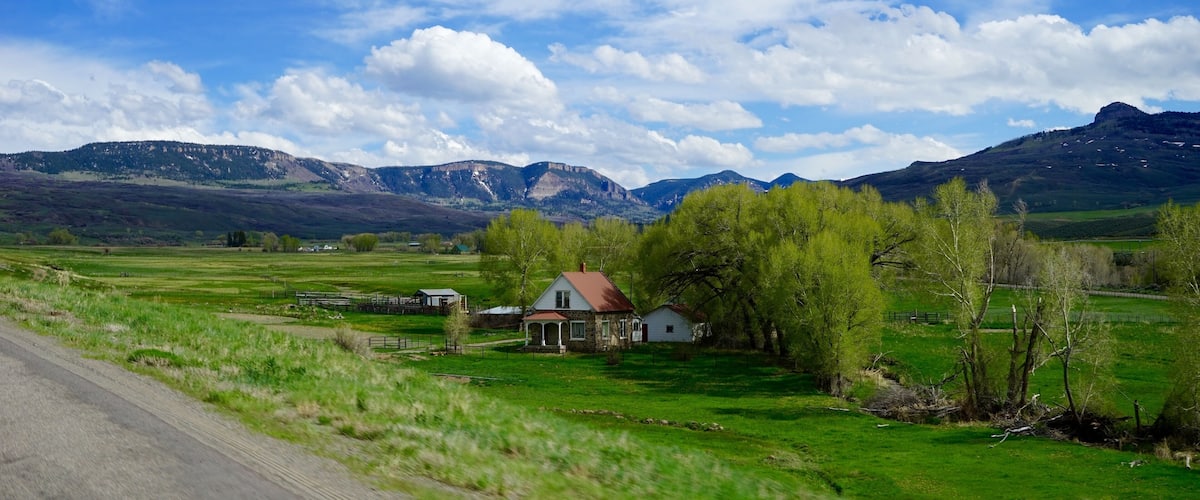 A rural landscape with a house, green fields, and mountains in the background.