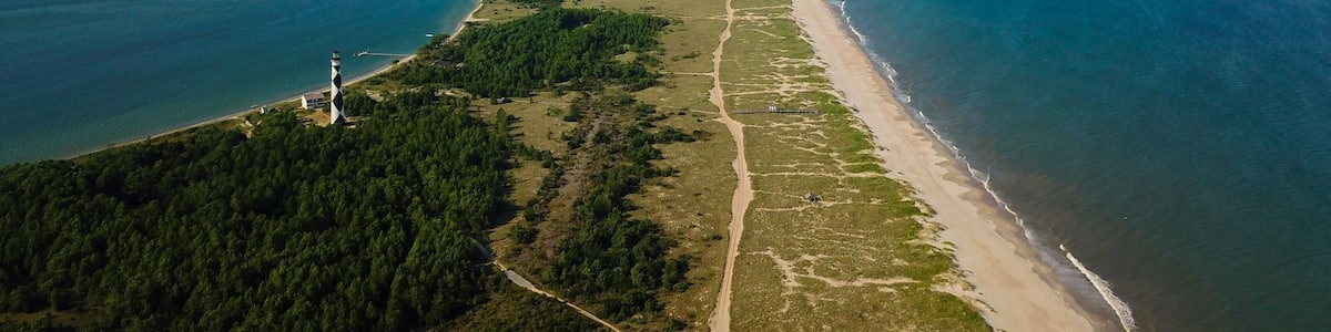 A coastal landscape with a long sandy beach, dense green forest, and a lighthouse.
