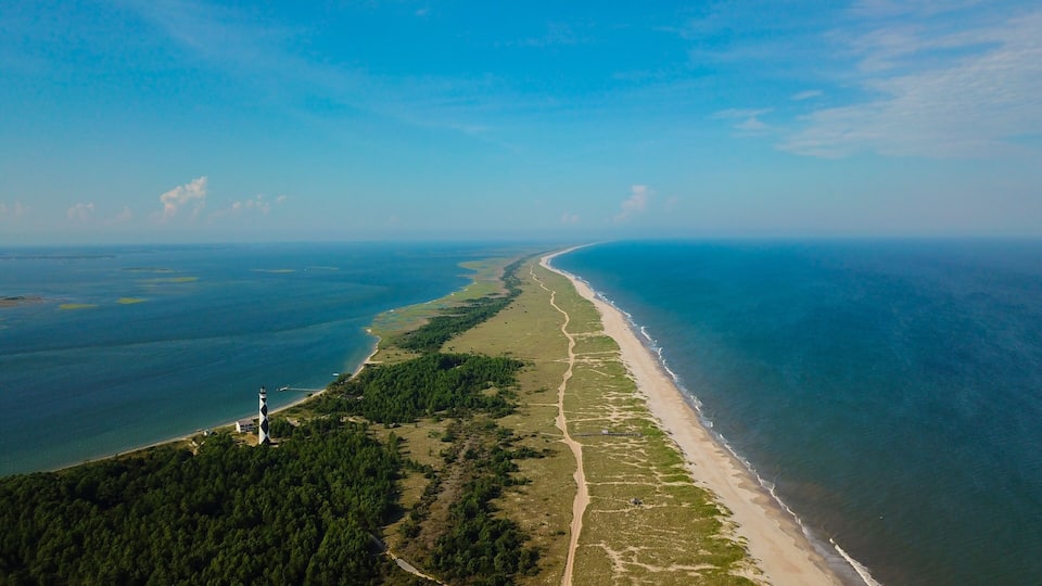 A coastal landscape with a long sandy beach, dense green forest, and a lighthouse.