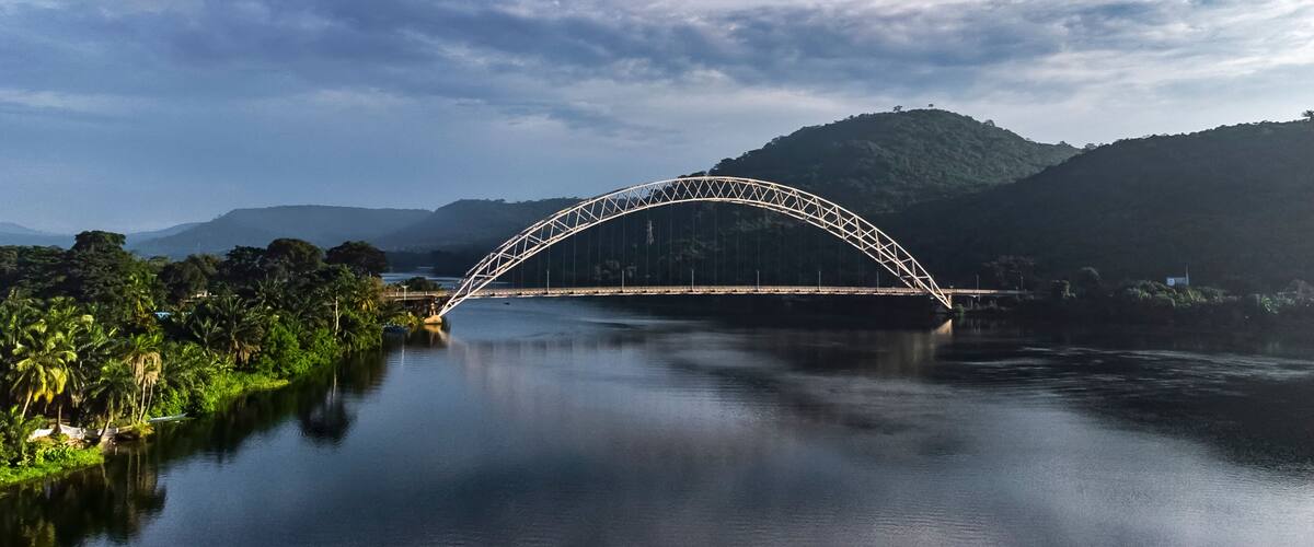 Aerial view of the beautiful Adomi Bridge over a tranquil river surrounded by lush hills and a serene sky, Atimpoku, Ghana.