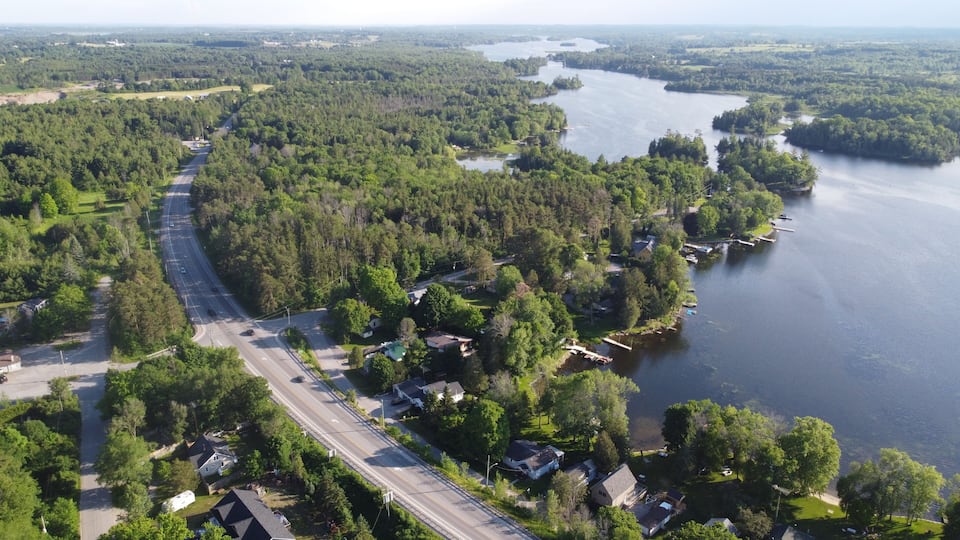 An aerial view of a residential area with a road running alongside a river, surrounded by dense forests and small houses.