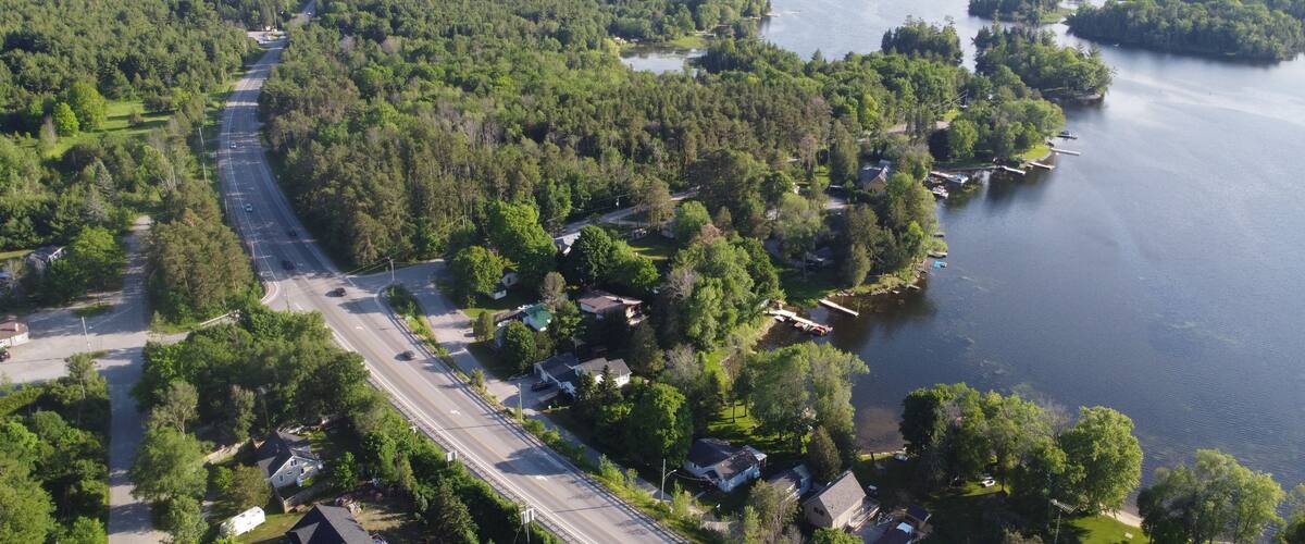 An aerial view of a residential area with a road running alongside a river, surrounded by dense forests and small houses.