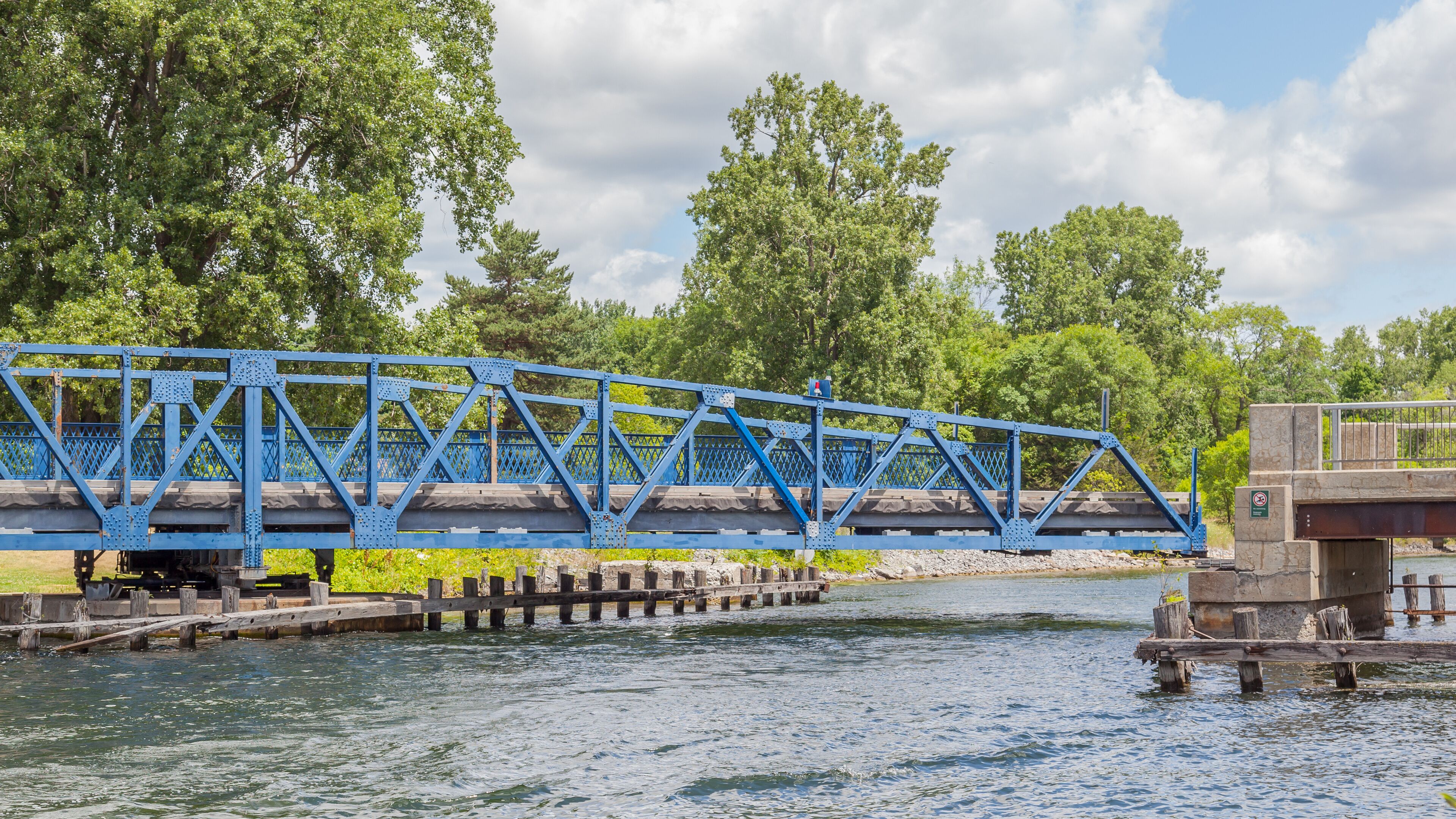 A blue steel bridge over a river with trees in the background.