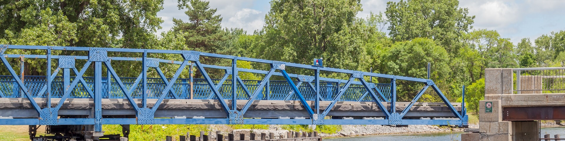 A blue steel bridge over a river with trees in the background.