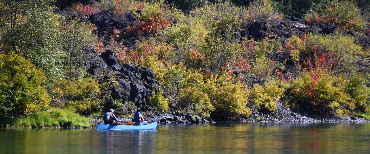 A calm lake with a small blue boat carrying two people. The surrounding area is filled with colorful autumn foliage.