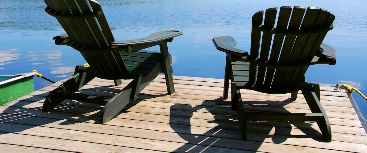 Two adirondack wooden chairs on dock facing a blue lake with clouds reflections