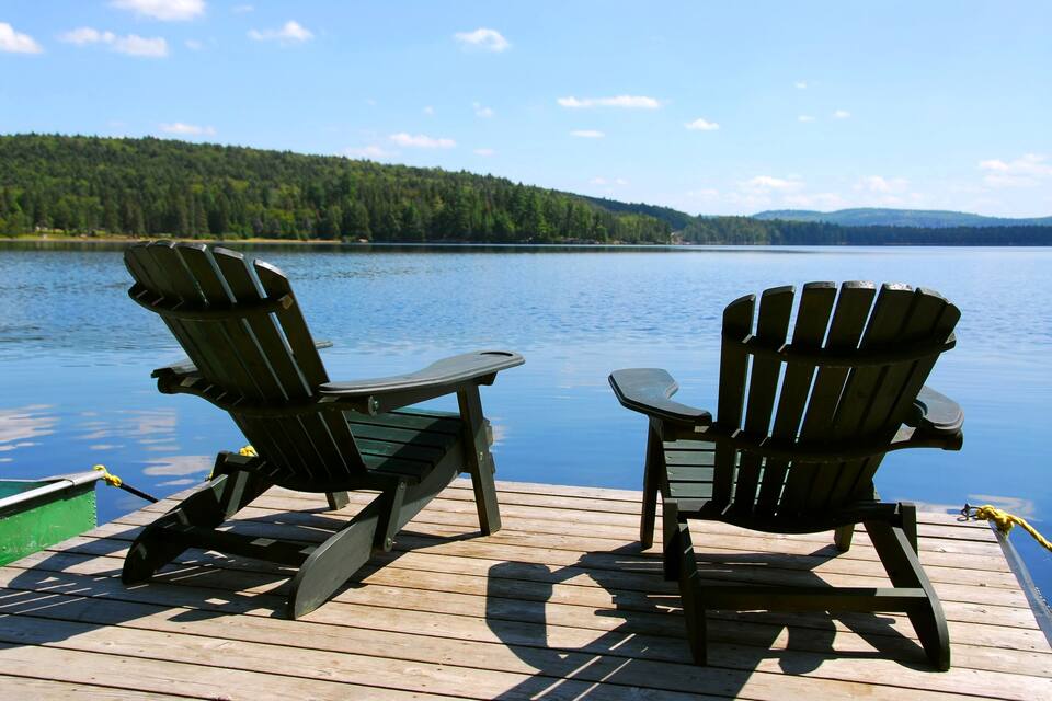 Two Adirondack chairs on a wooden dock overlooking a calm lake with a forested hill in the background.