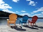 Three Adirondack chairs on a wooden dock by a lake with a forested hill in the background.